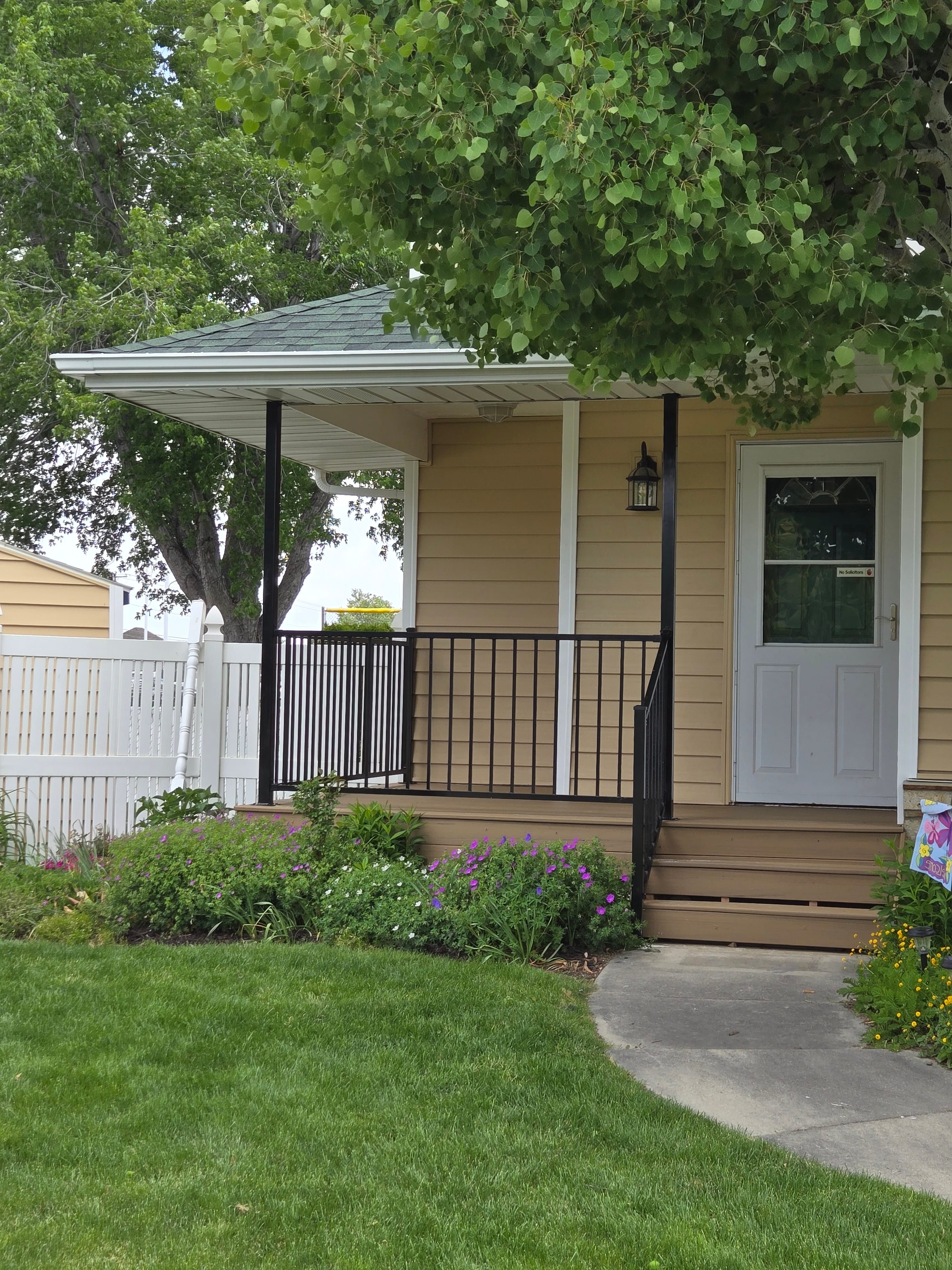 Front porch with wood-toned decking, steps, and black metal railing beside a garden